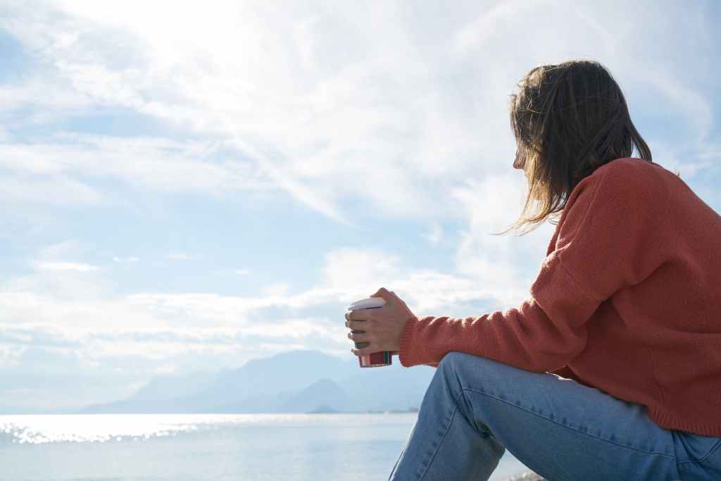 Young lady in jumper and jeans sitting pondering with paper cup of drink in hands and a backdrop of see, clouds and a vauge outline of hills on the other side of a bay